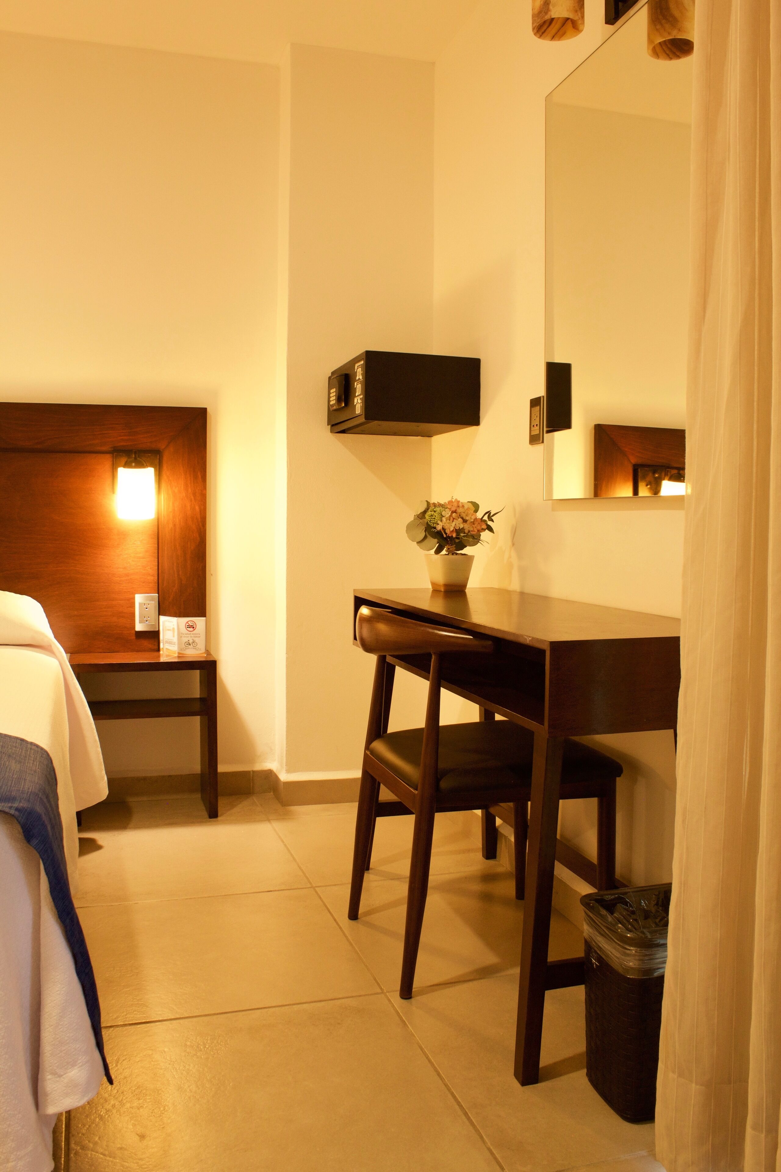A hotel room with a bed, a desk, a chair, a trash bin, a wall-mounted TV, and a small table with a vase of flowers.