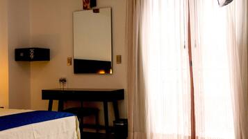 A hotel room with a bed, a desk, a chair, a flat-screen TV mounted on the wall, and a large window with sheer curtains.