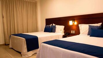 A hotel room with two beds, each with a blue bedspread and white linens. There is a wooden headboard with wall-mounted lights, a curtain on the window, and a telephone on the nightstand.