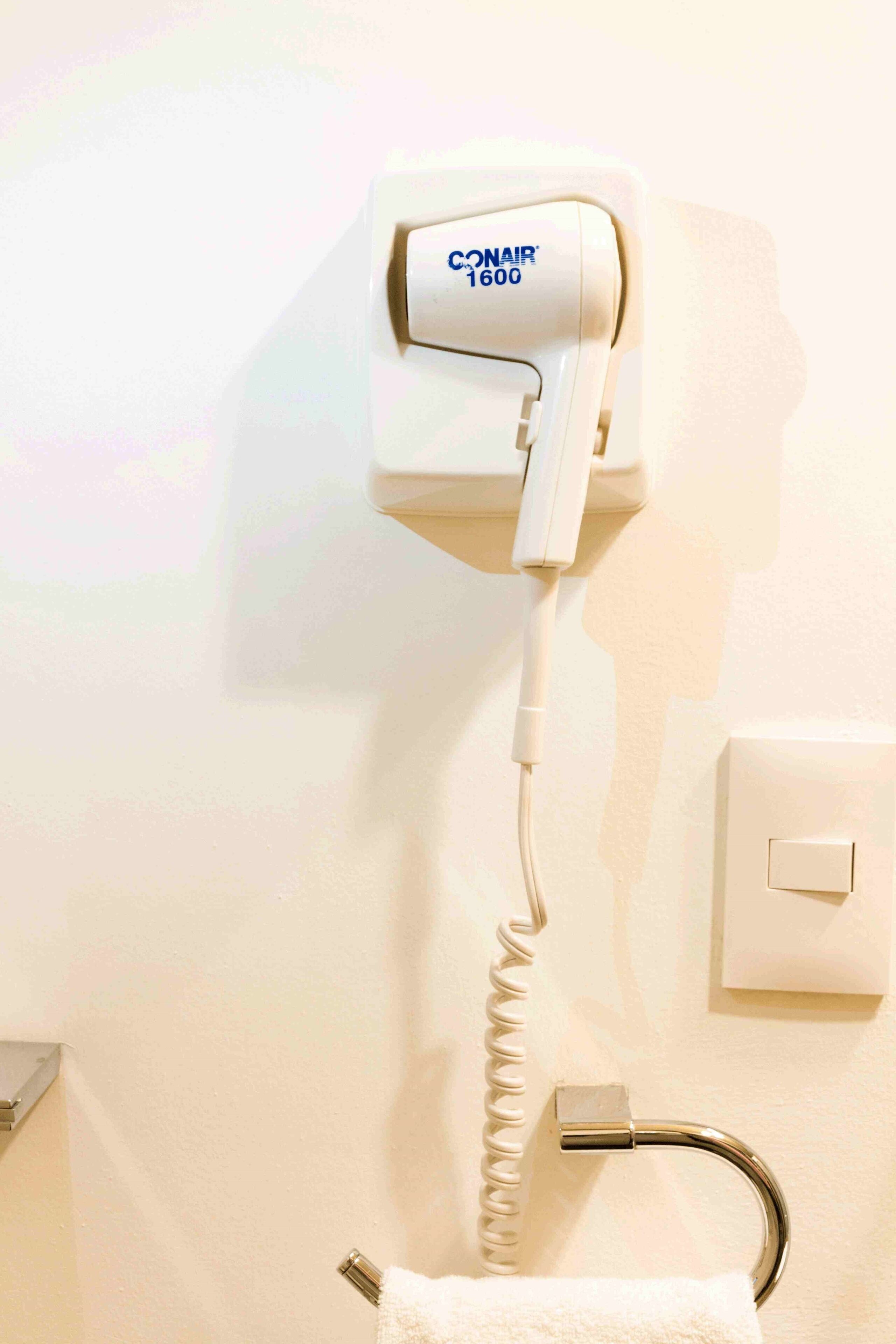 A hair dryer mounted on a wall with a coiled cord, a towel rack, and a light switch.