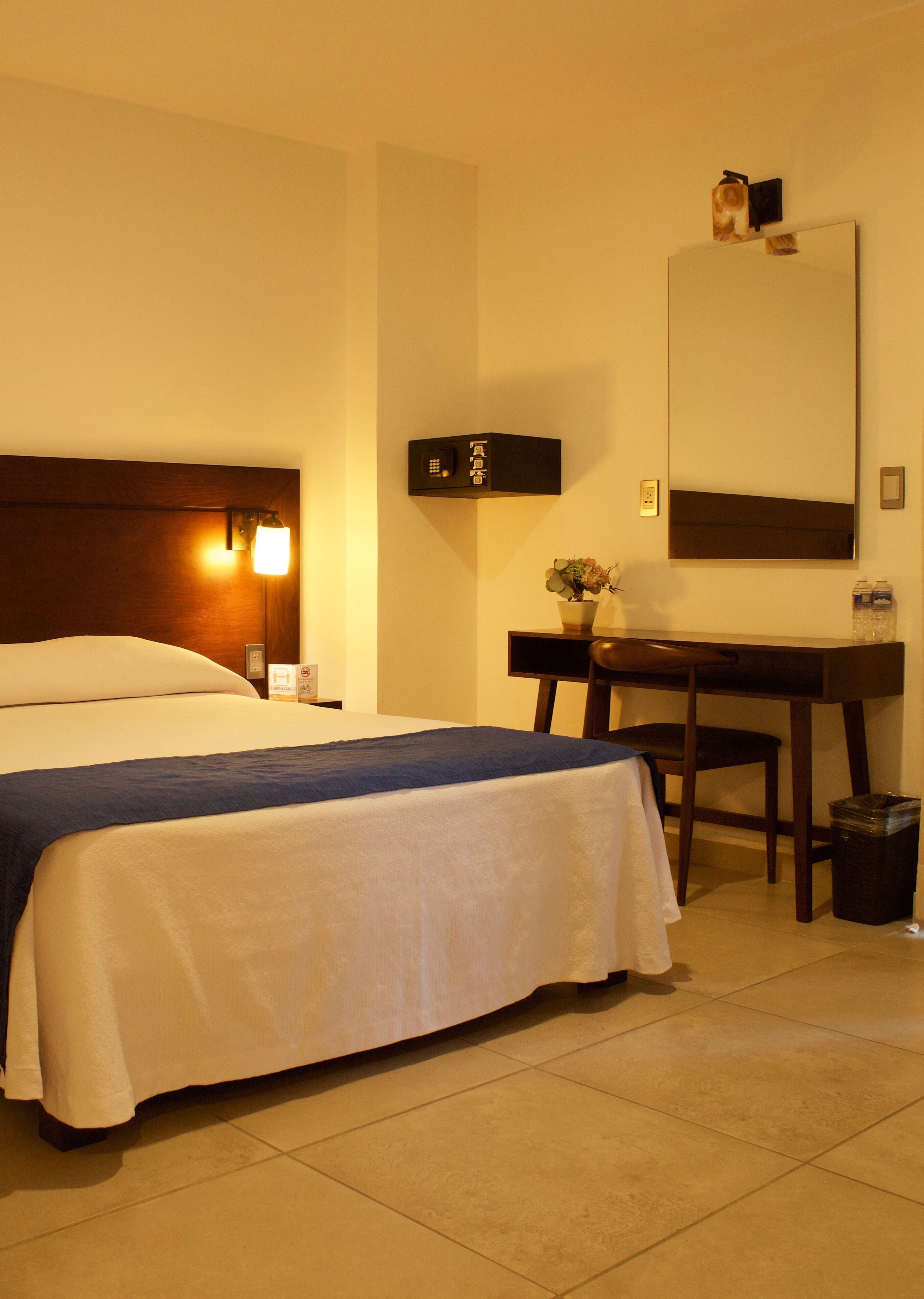 A hotel room with a bed, bedside table, desk, chair, and a trash bin.