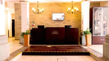A hotel reception area with a dark reception desk, two chandeliers, a flat-screen TV, and a large potted plant.