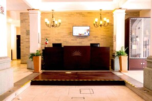 A hotel reception area with a dark reception desk, two chandeliers, a flat-screen TV, and a large potted plant. - Hotel Real Alma  (Oaxaca)