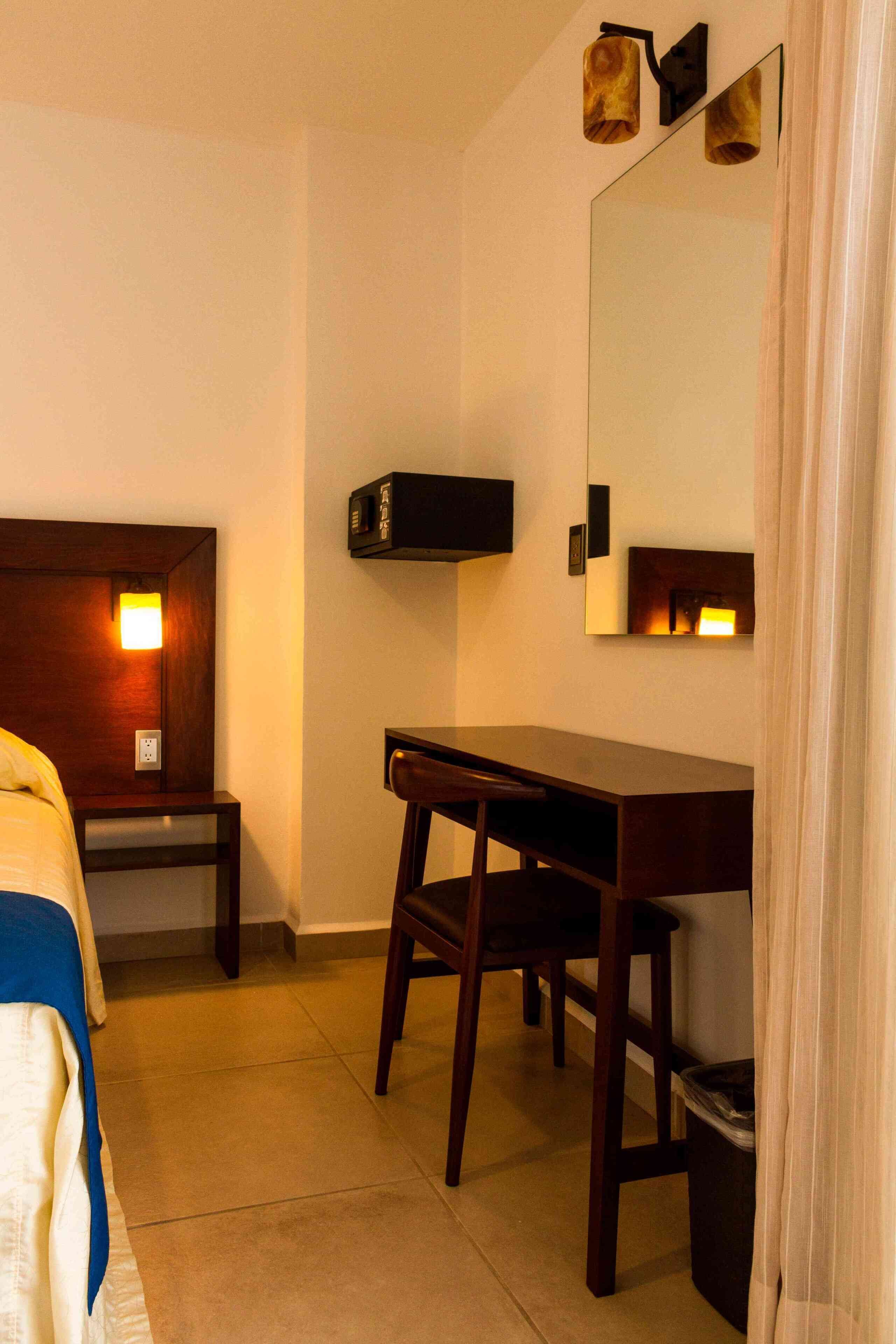 A hotel room with a bed, a desk, a chair, a trash bin, and a mirror.