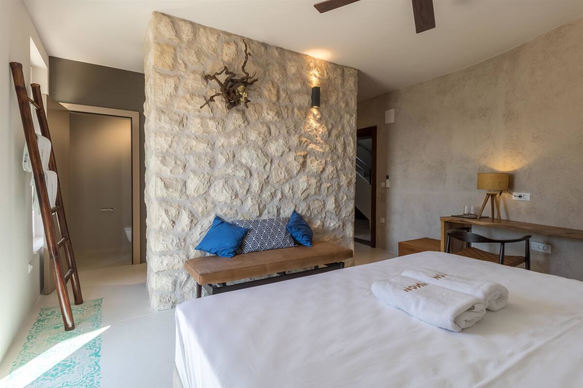 A hotel room with a stone accent wall, a wooden bench with blue cushions, a desk with a lamp, and a neatly made bed with towels.