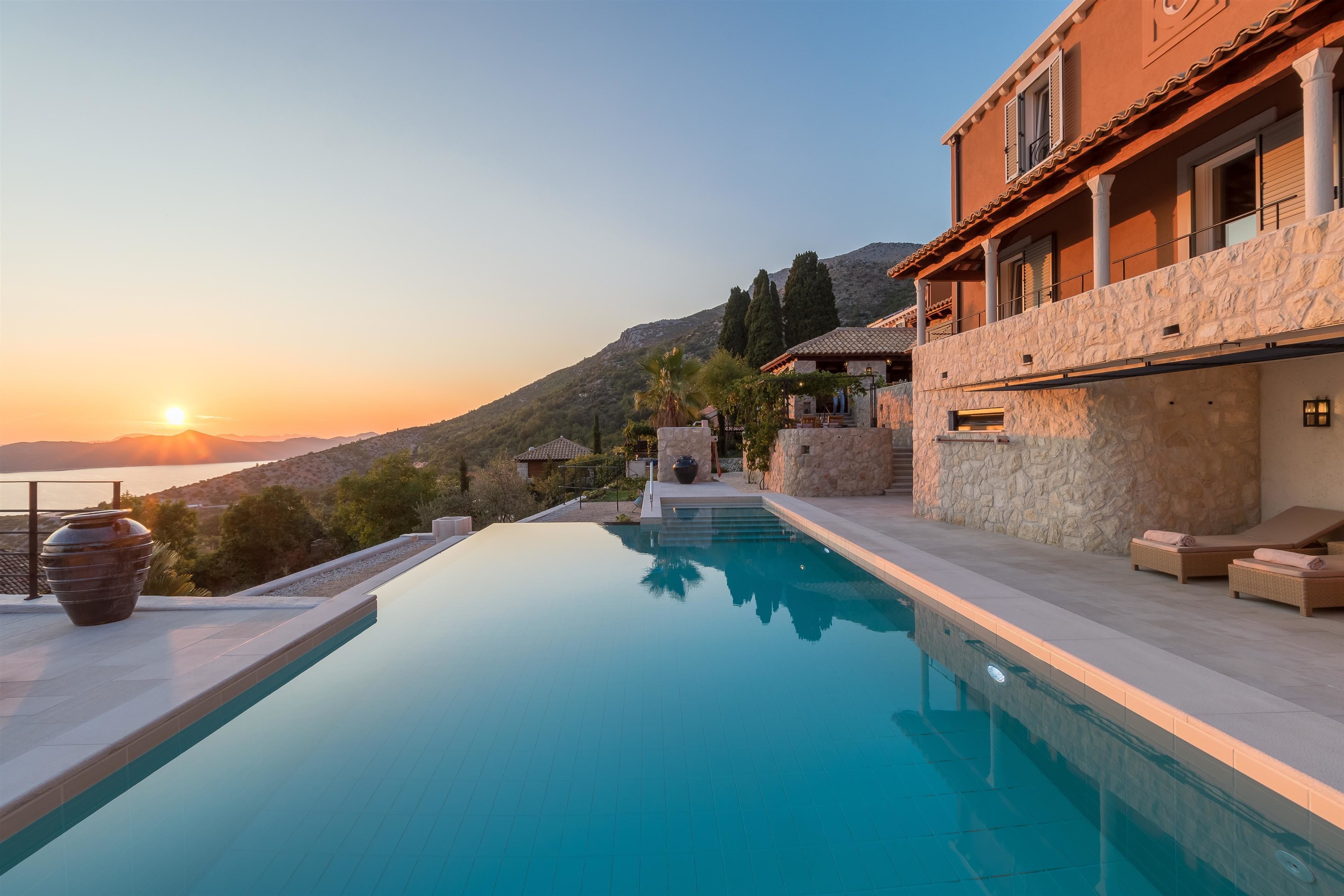 Vista de la piscina de una villa de estilo mediterráneo con fachada de piedra, un balcón con sillones de descanso y la vista del atardecer sobre el mar.