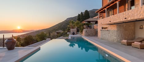 Vista da piscina de uma villa em estilo mediterrâneo, com fachada de pedra, varanda com espreguiçadeiras e pôr do sol sobre o mar.