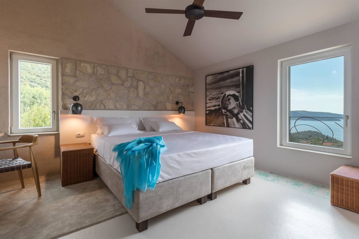 A bedroom with a bed, a desk, a chair, a ceiling fan, a stone wall, a painting, and a view of the sea.