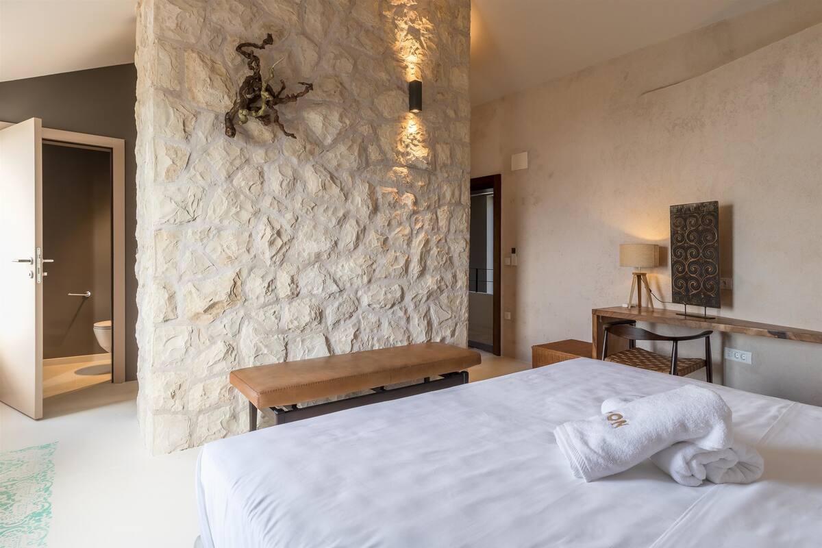 A bedroom with a stone accent wall, a bed with a white duvet and a towel, a bench, a bedside table with a lamp, and a mirror on the wall.