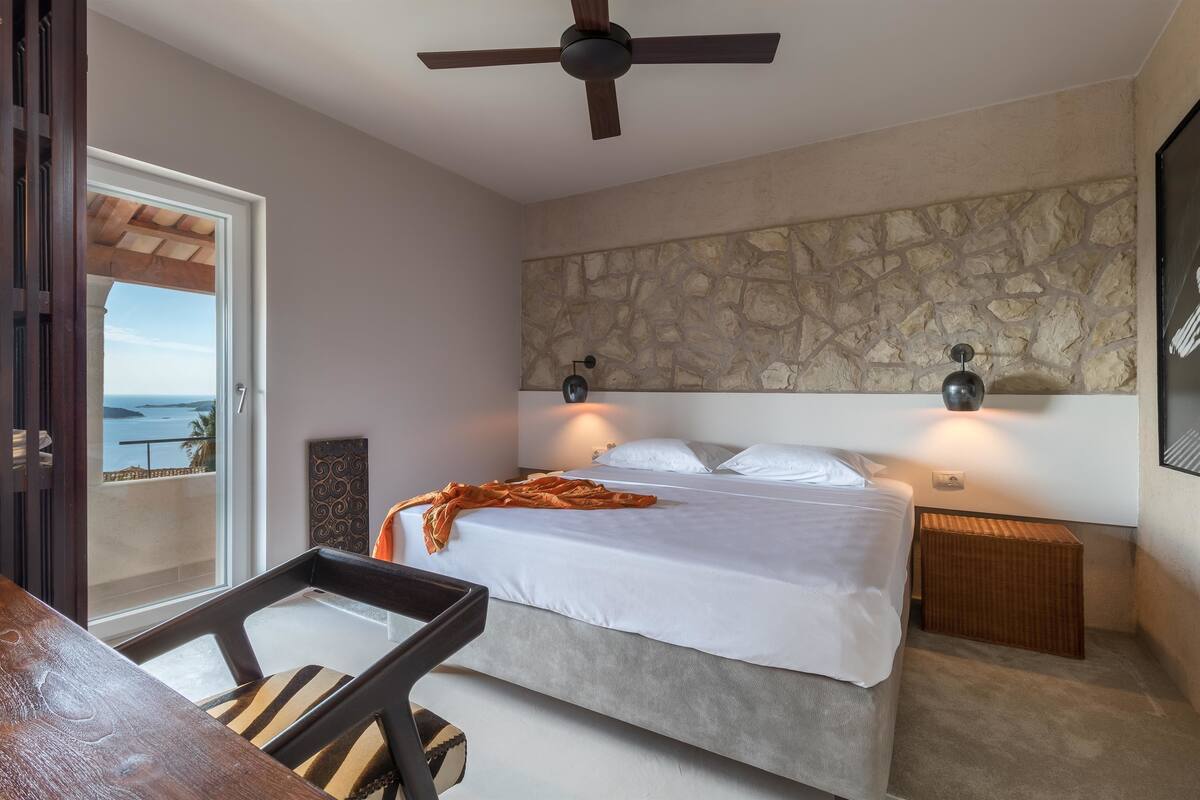 A bedroom with a bed, a ceiling fan, a stone wall, and a view of the ocean.