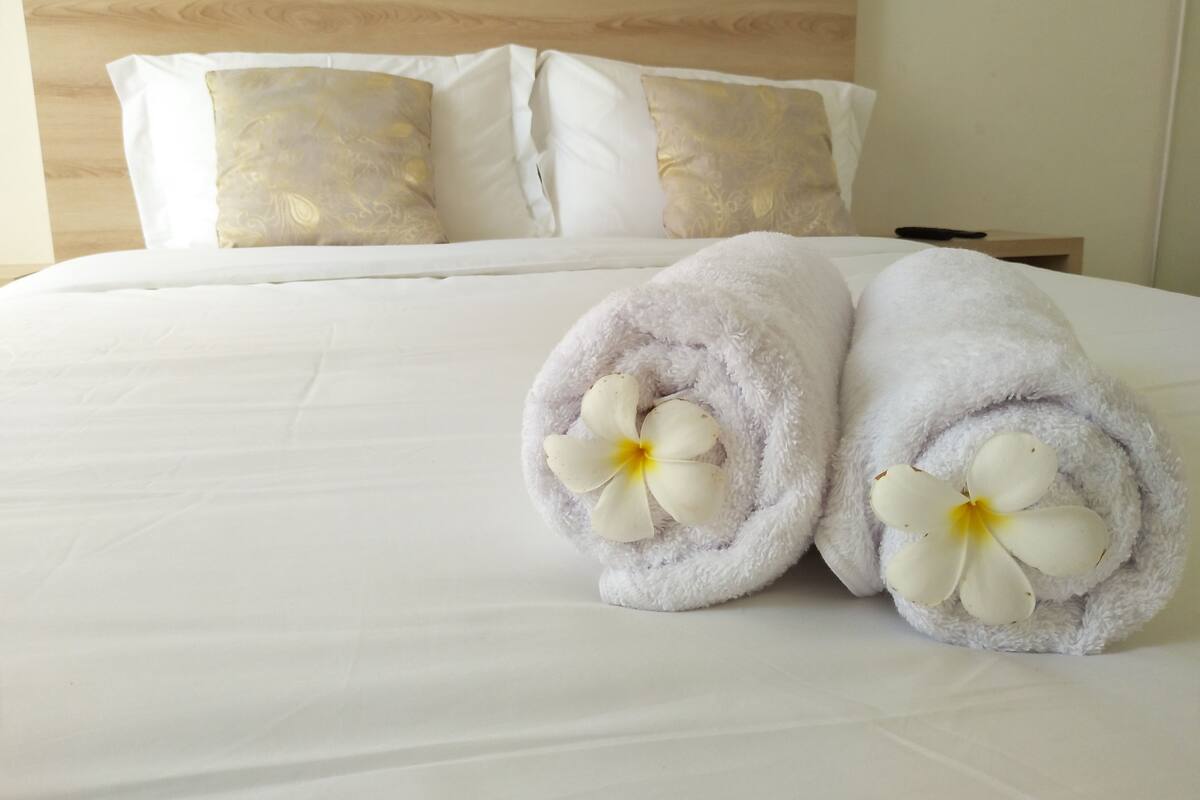 Un lit bien fait, avec du linge de lit blanc et deux serviettes enroulées, ornées de fleurs blanches.
