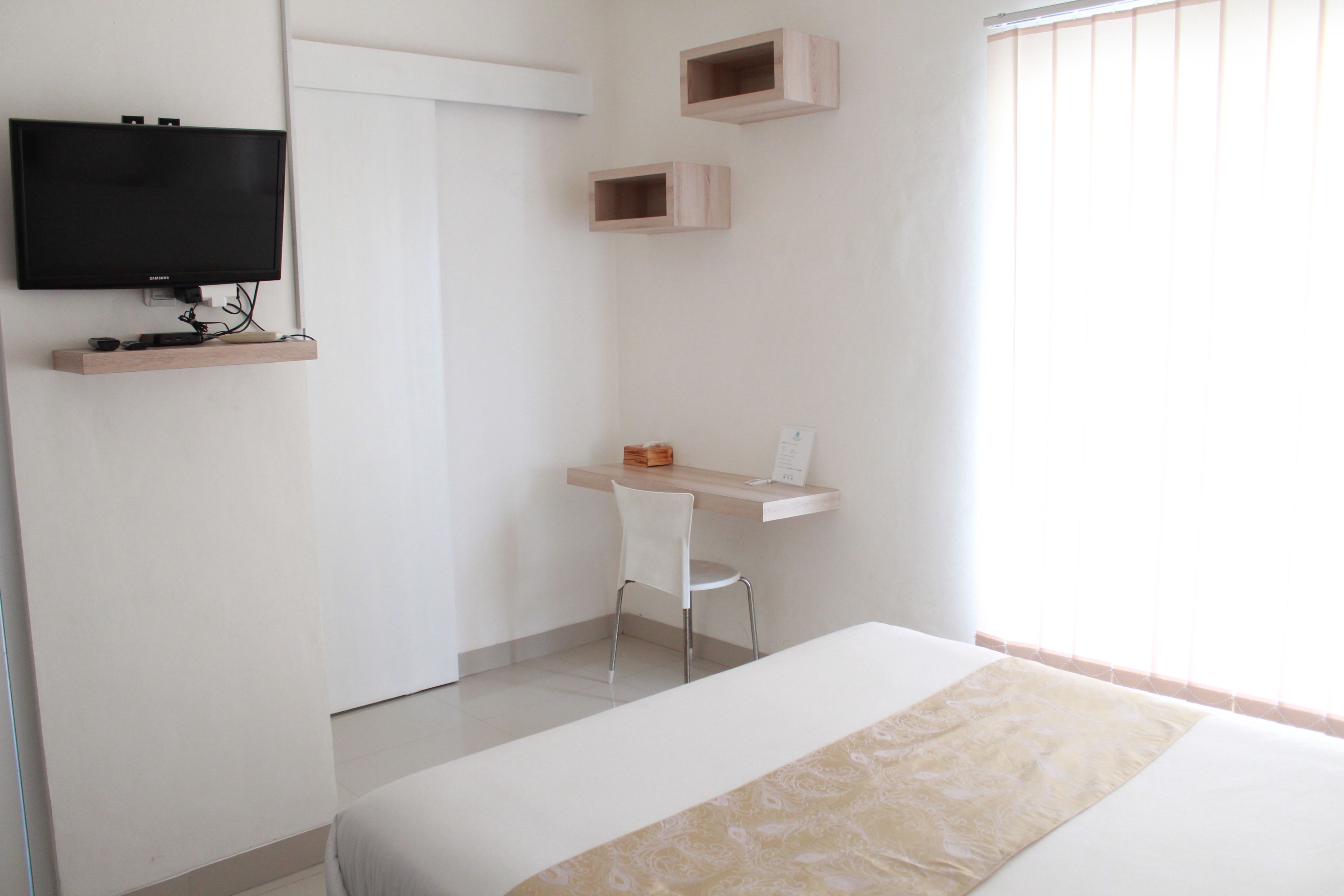 A bedroom with a bed, a desk, a chair, a TV, and a window with blinds.