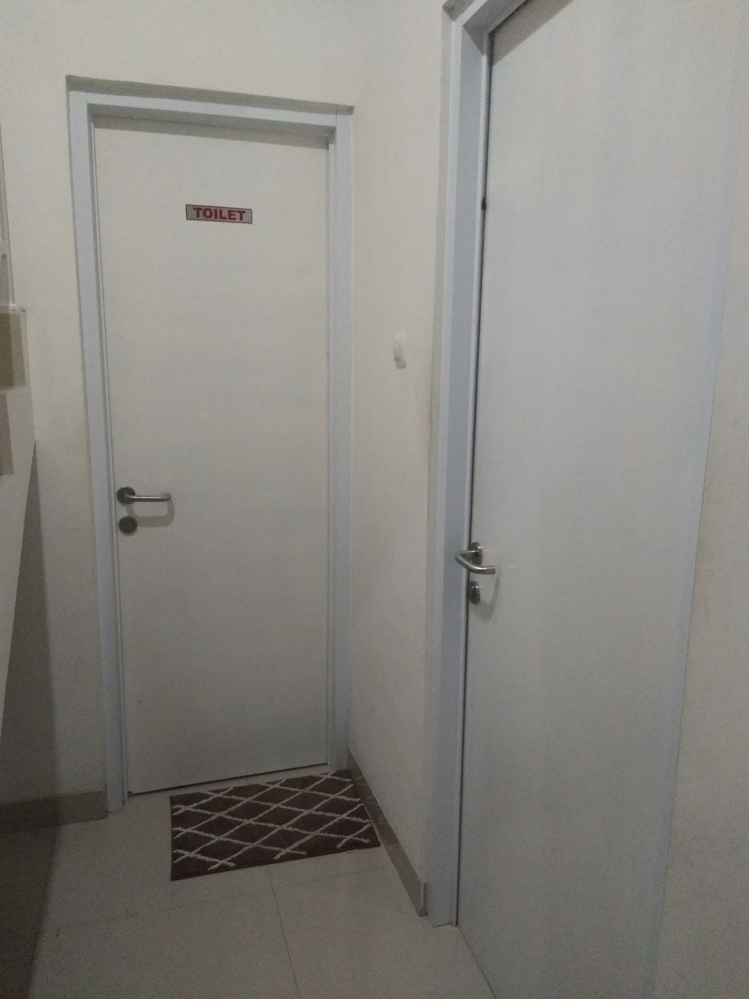 Een witte deur met een "TOILET" bord, een metalen hendel en een klein matje onderaan.