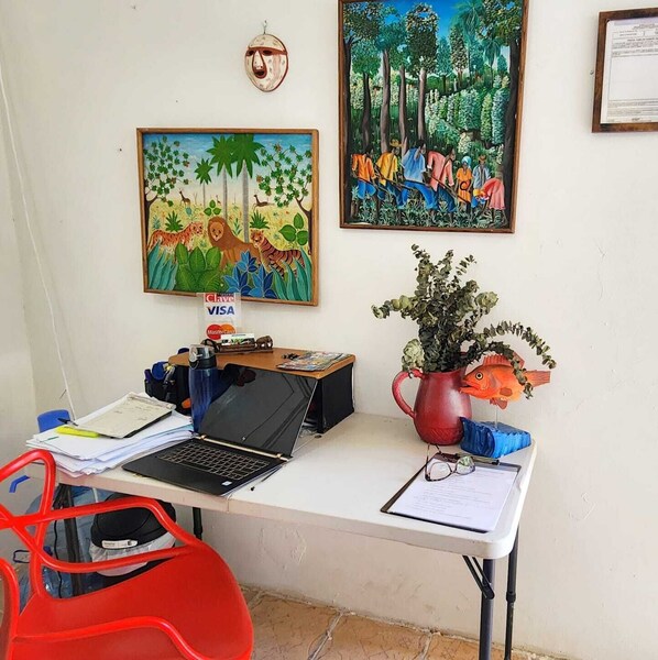 A desk with a laptop, papers, and a red chair. There are two paintings on the wall and a wooden mask hanging above the desk. - Cerrito Tropical (Taboga)