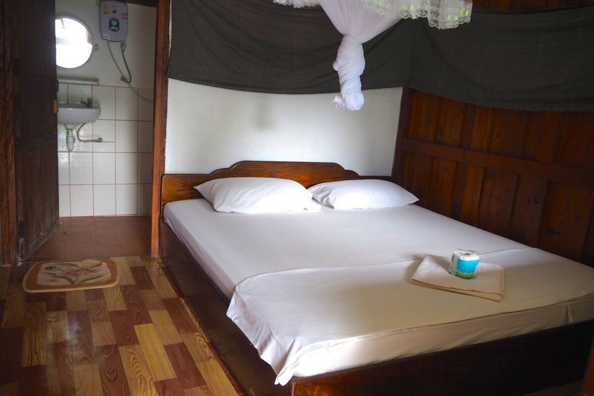 Una cama de madera con ropa de cama blanca, una mesita de noche con un objeto azul y un suelo de madera.