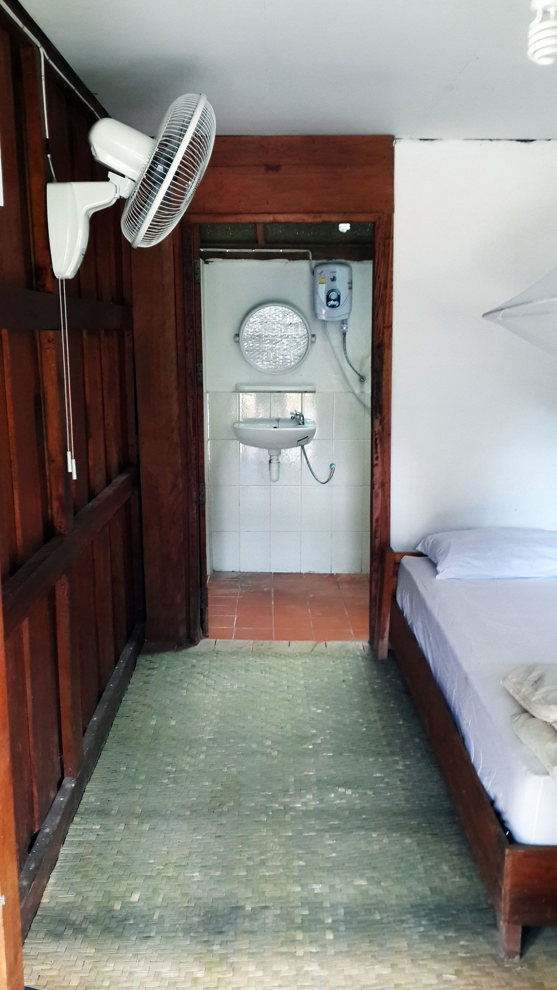 A small, narrow room with a wooden floor, a bed with a white mattress, a white sink, and a wall-mounted fan.