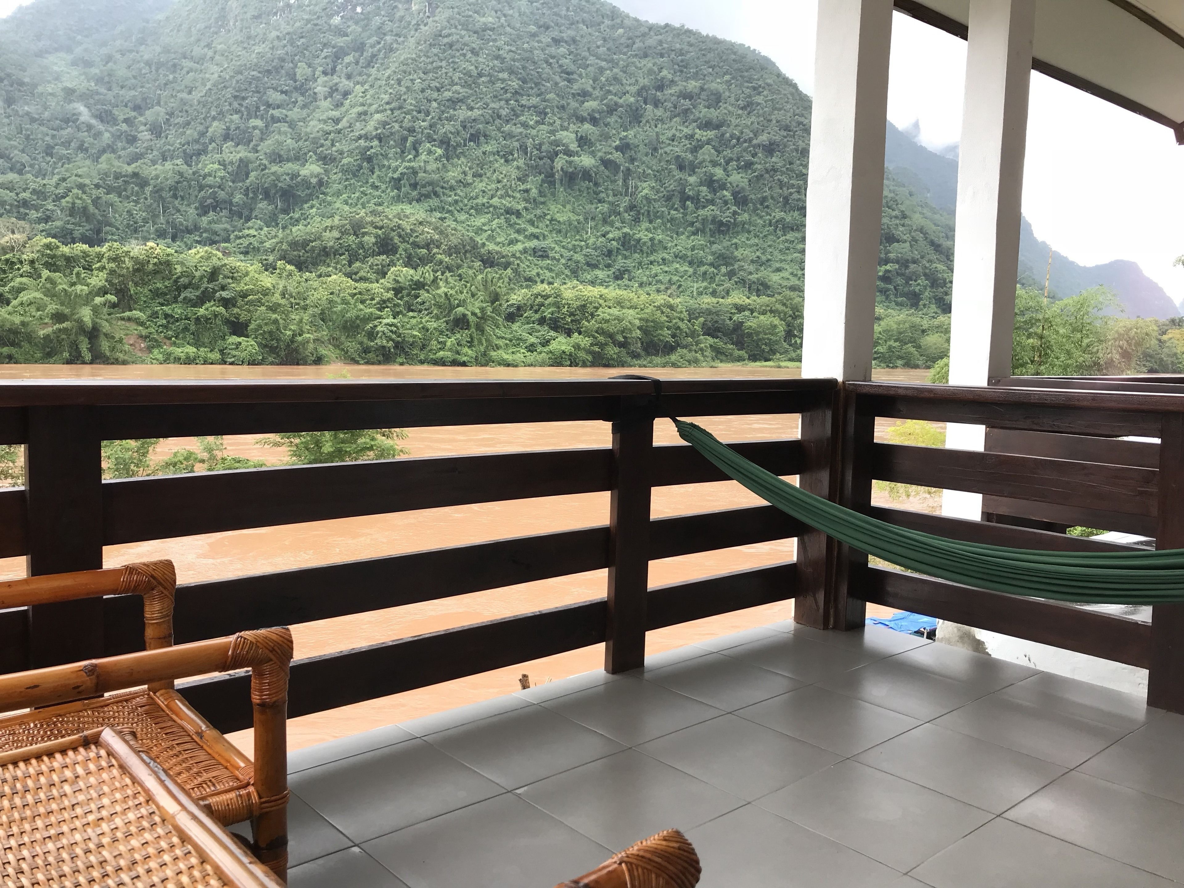Uma varanda com vista para uma montanha arborizada, uma rede e móveis de vime.