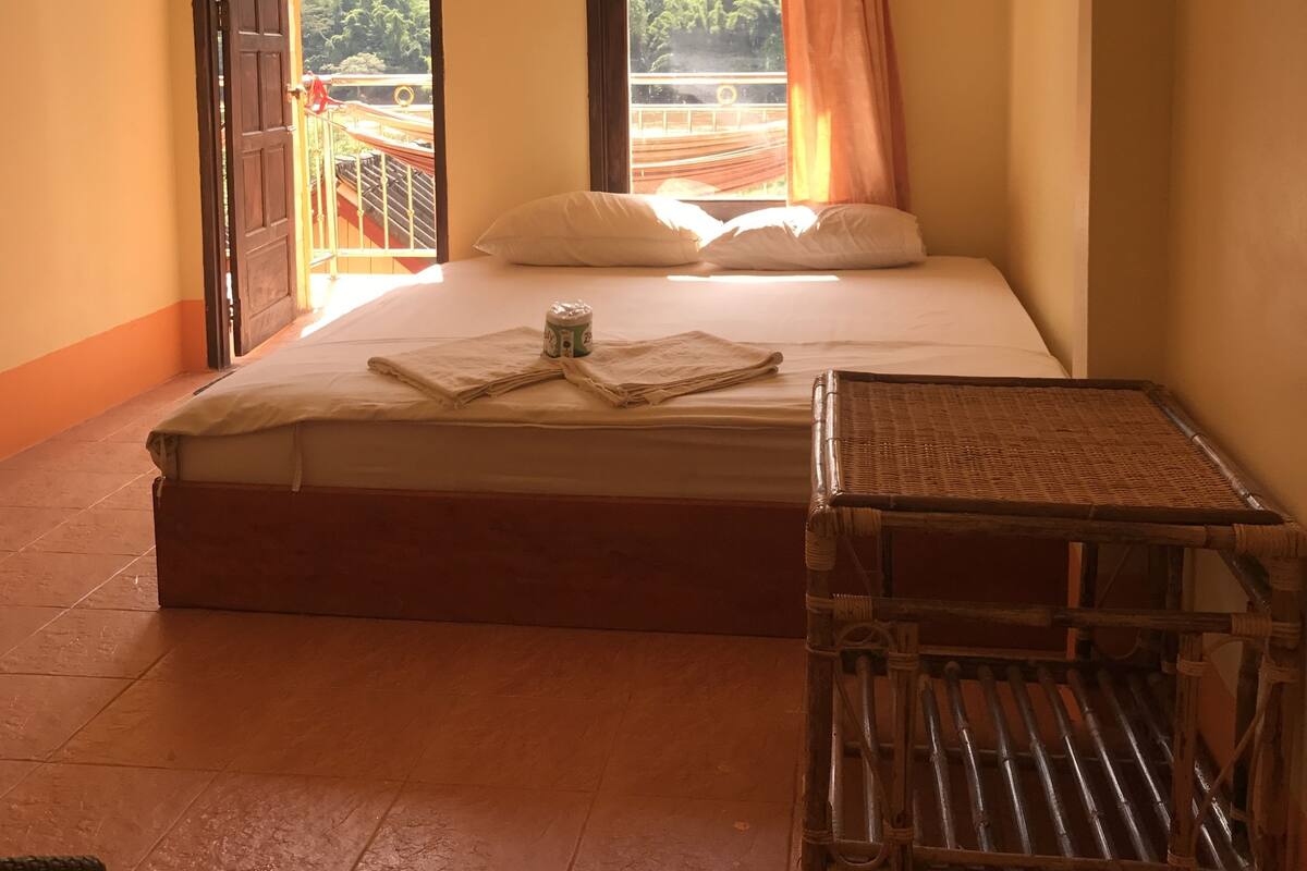 Un dormitorio con cama, ventana con vistas a la vegetación, ventilador de techo y una pequeña mesa.