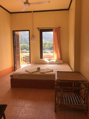 A bedroom with a bed, a window with a view of greenery, a ceiling fan, and a small table.