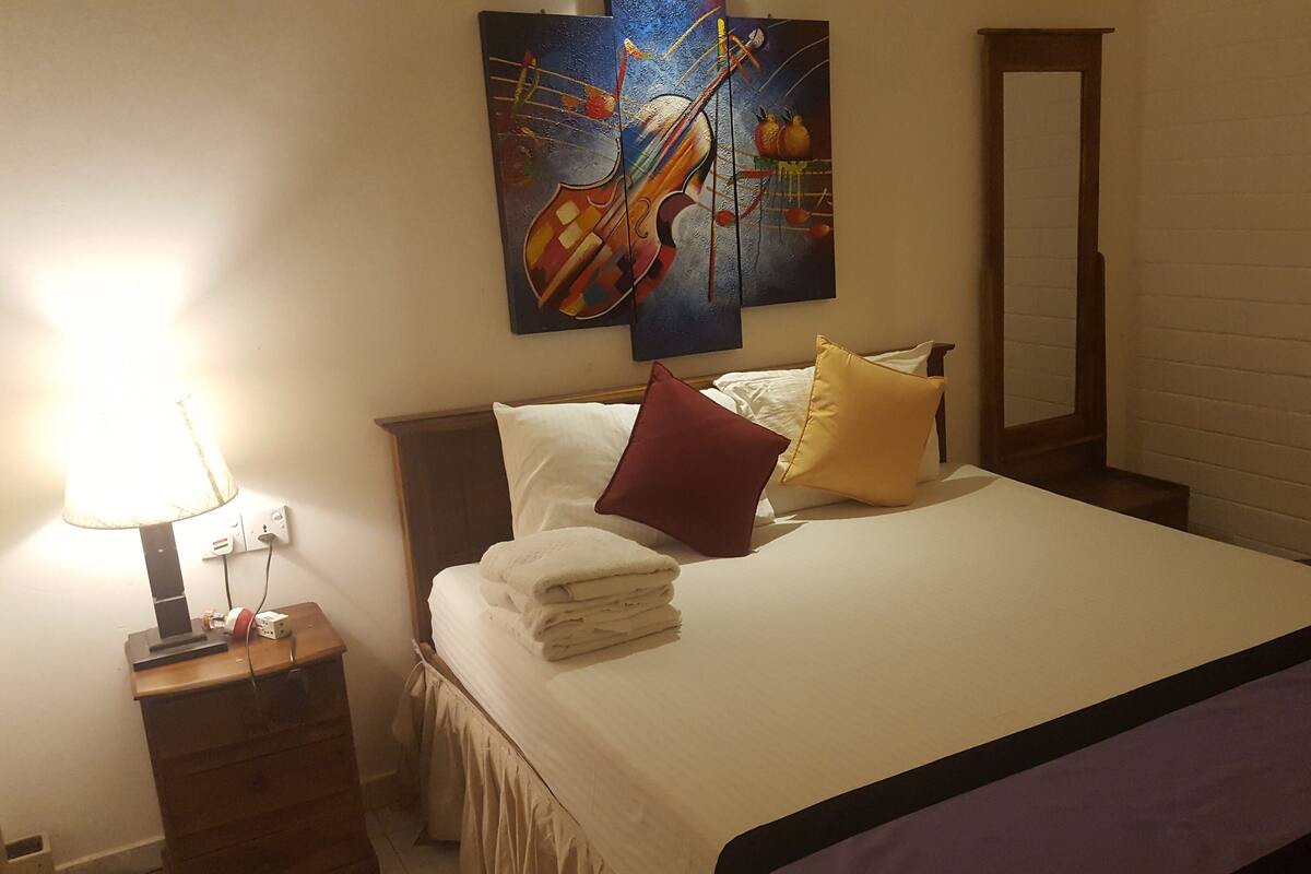 A neatly made bed with white linens and a purple accent, a bedside lamp, and a wall-mounted painting of a guitar.