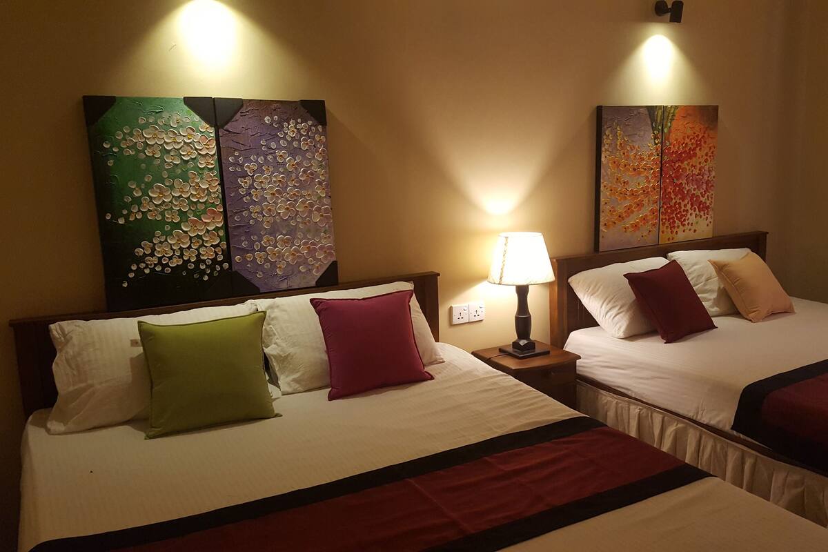 Two adjacent hotel beds with white linens, green and red pillows, and a lamp between them.