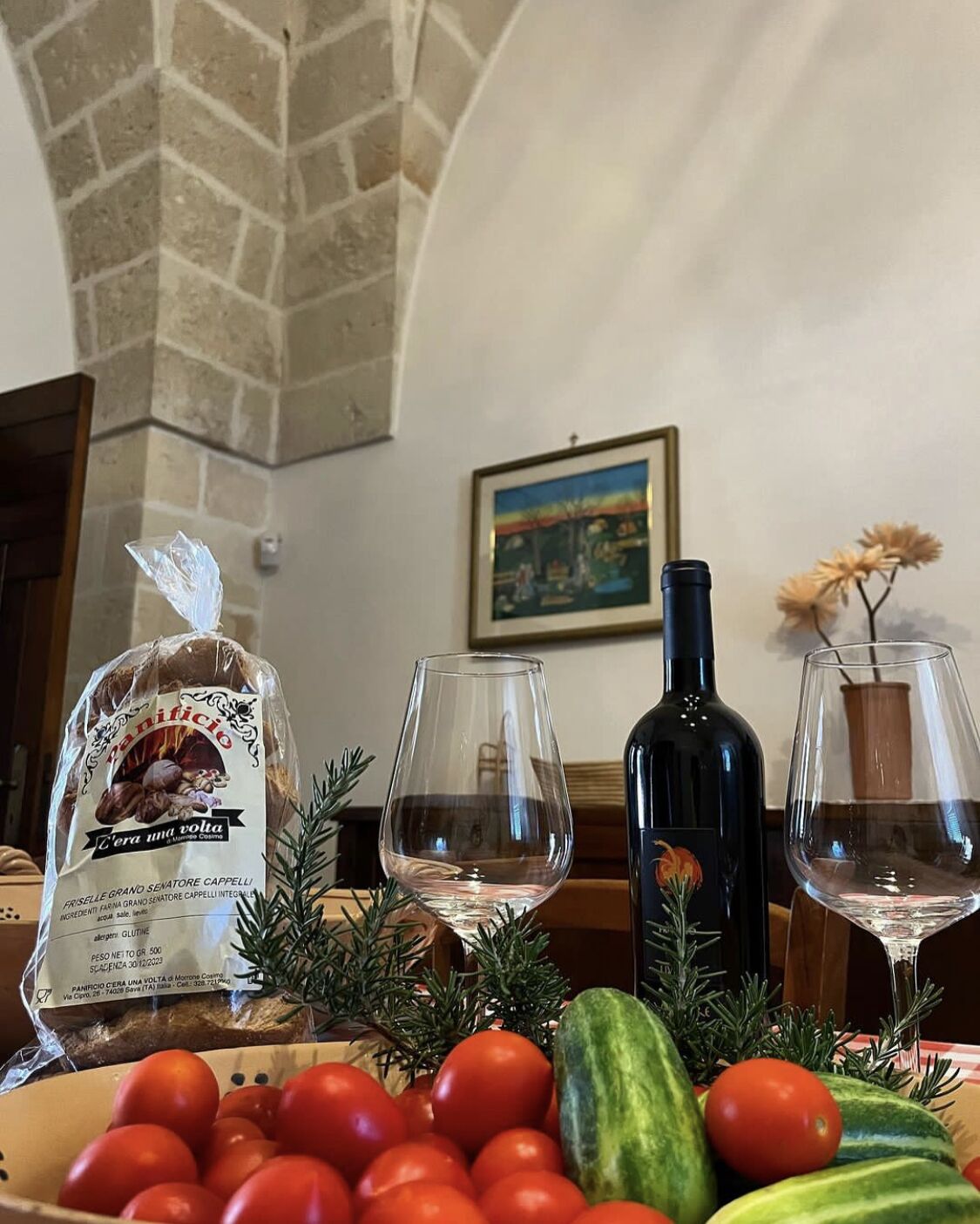 Un tavolo con pane, pomodori, cetrioli e una bottiglia di vino in una stanza con pareti in pietra e un quadro appeso.