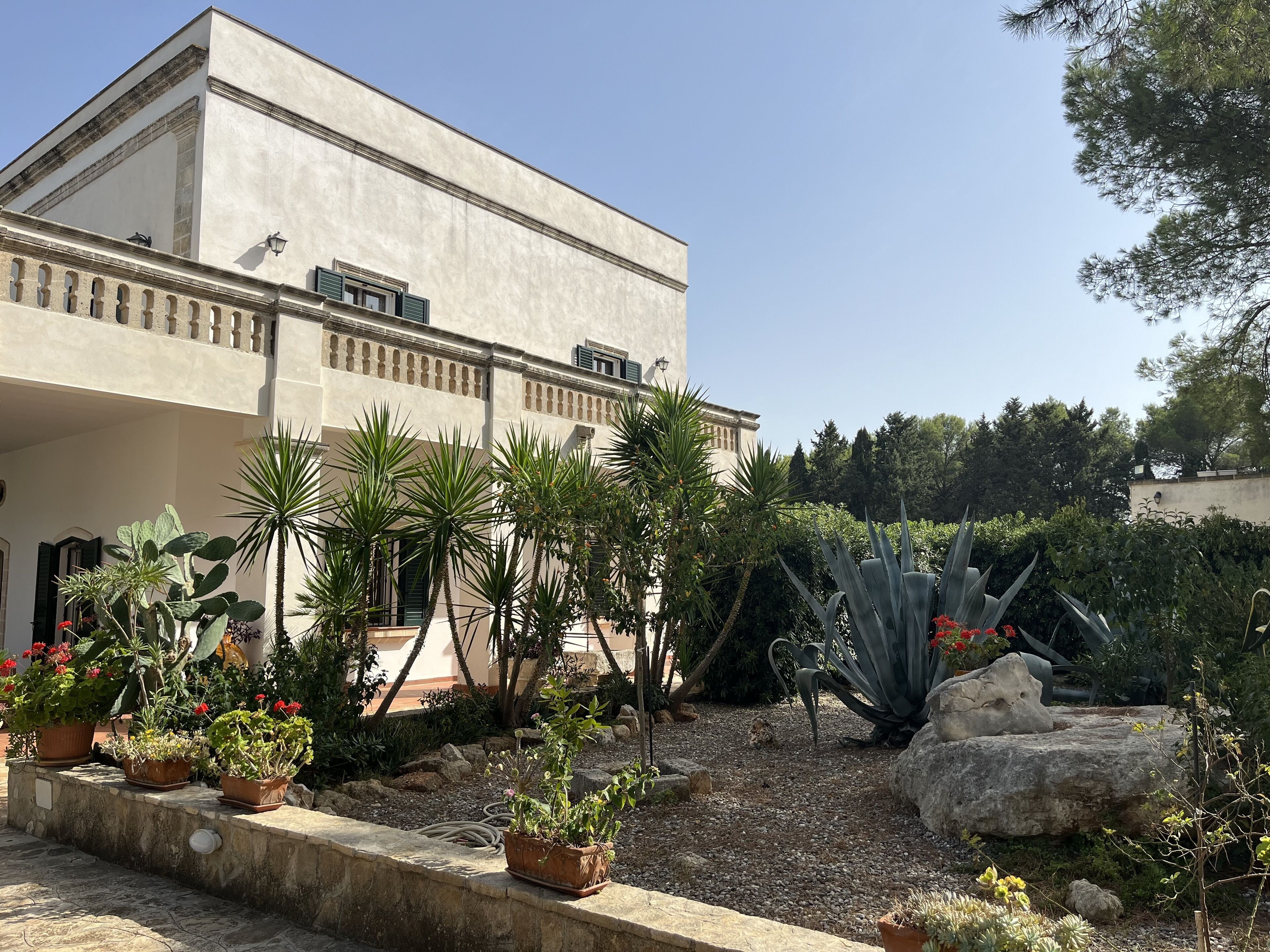 A building with a flat roof and a balcony with a railing. There are potted plants and a rock garden in front of the building.
