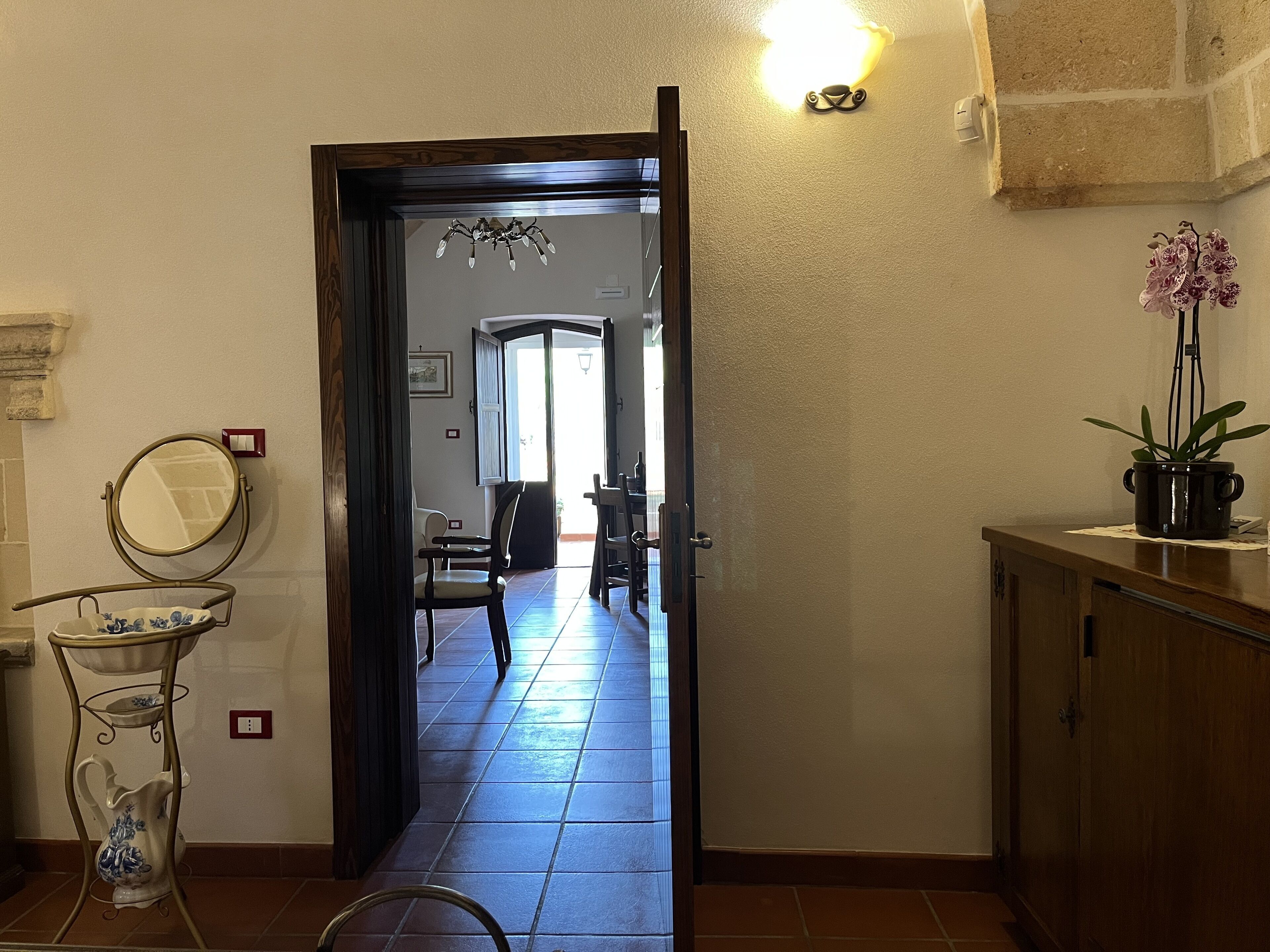 Um corredor com uma porta de madeira que dá para uma sala de jantar, uma pequena mesa com um vaso e uma planta em um balcão.