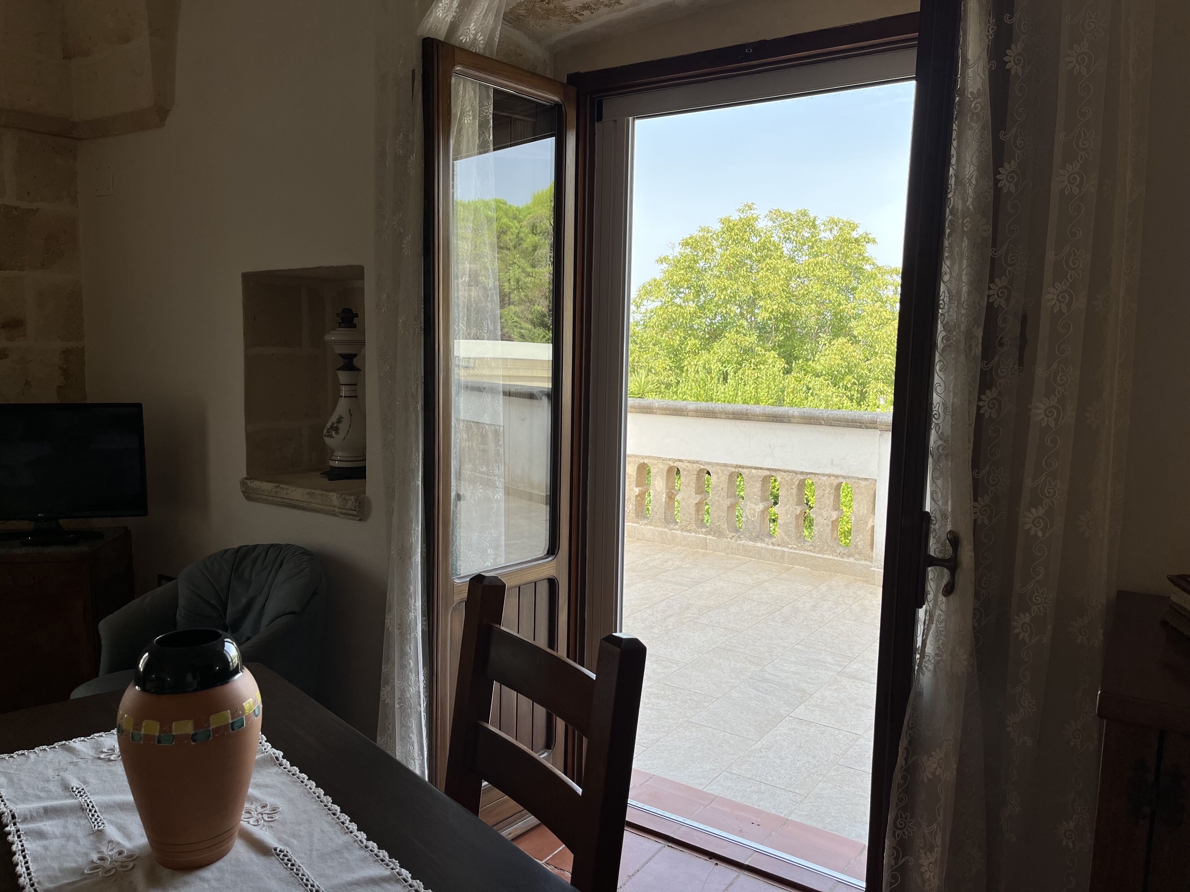 A room with a table, a chair, a vase, a television, and a door leading to an outdoor balcony with a view of trees and a railing.