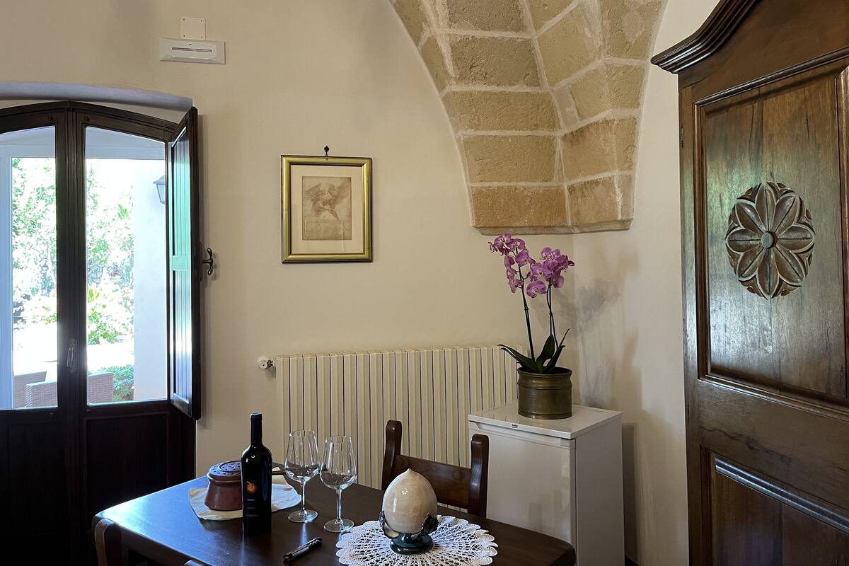 A dining area with a table set for two, a wine bottle, and two glasses. There is a potted orchid on the table, a framed picture on the wall, and a radiator below.