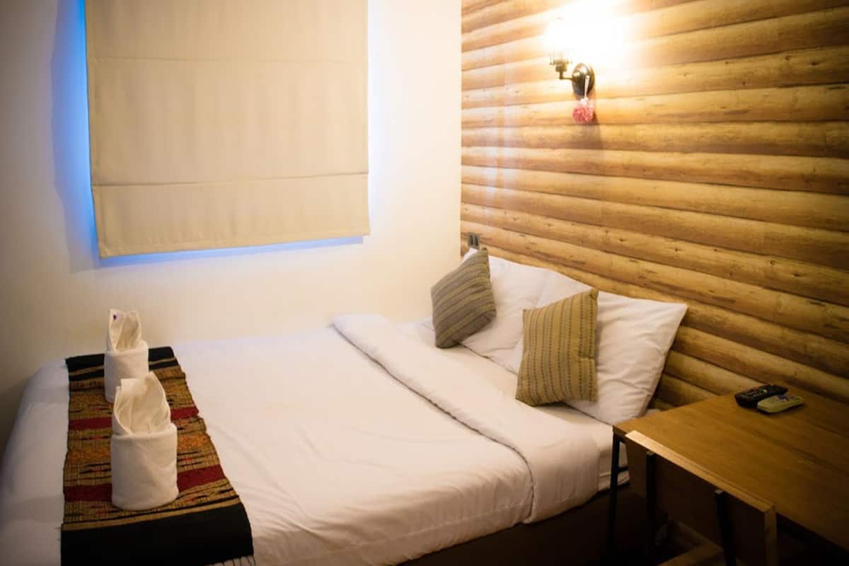 A single bed with a white comforter, two pillows, a blanket, and a tissue. A wooden headboard and a small table with a remote control are also visible.