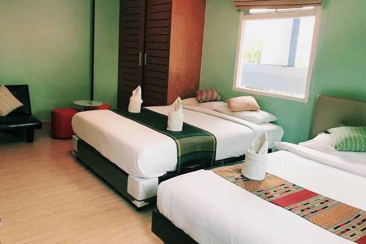 A hotel room with two beds, a red ottoman, a small round table, and a window with blinds.