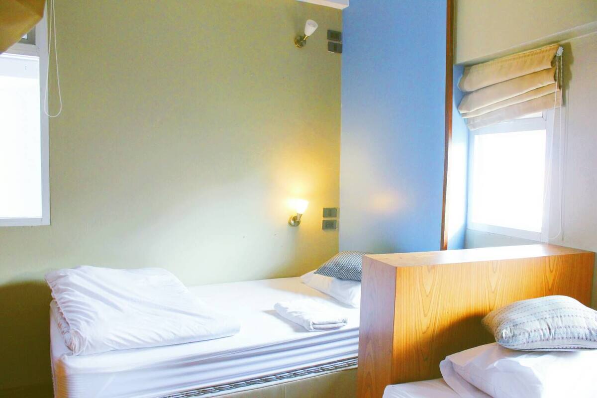 A hotel room with a bed, a wooden bedside table, a nightstand, a lamp, and a window with blinds.