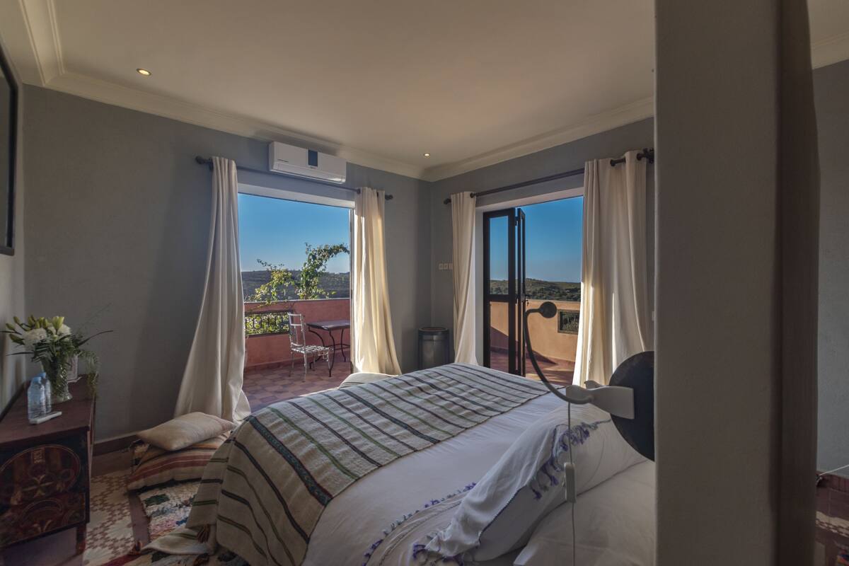 A bedroom with a large bed, a desk with a chair, and a view of the outdoors.