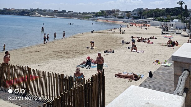 En strand med folk som solar och badar, ett trästaket och en klar himmel.