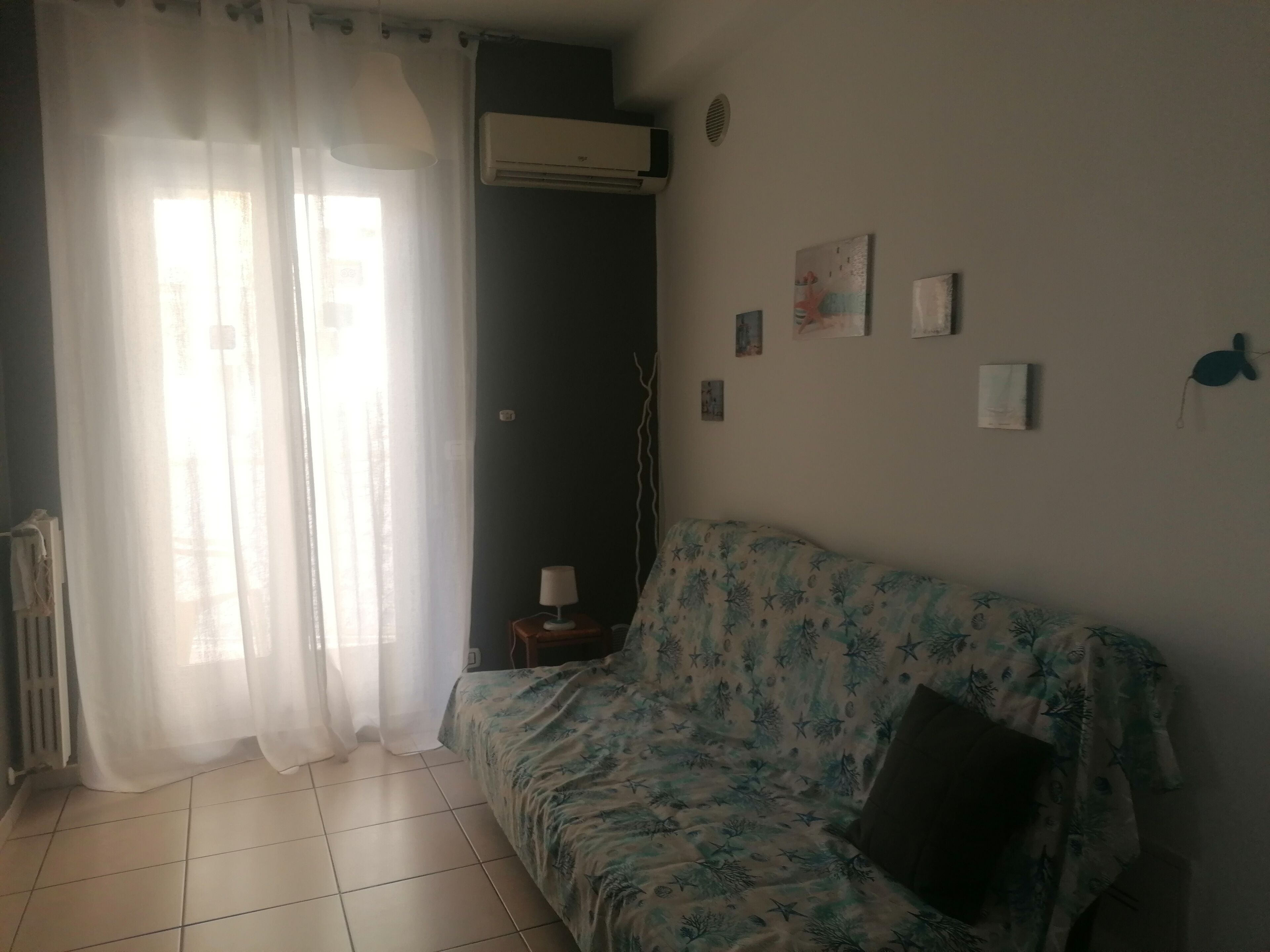 Quarto com cama, radiador, janela com cortinas leves e ar-condicionado fixado na parede.