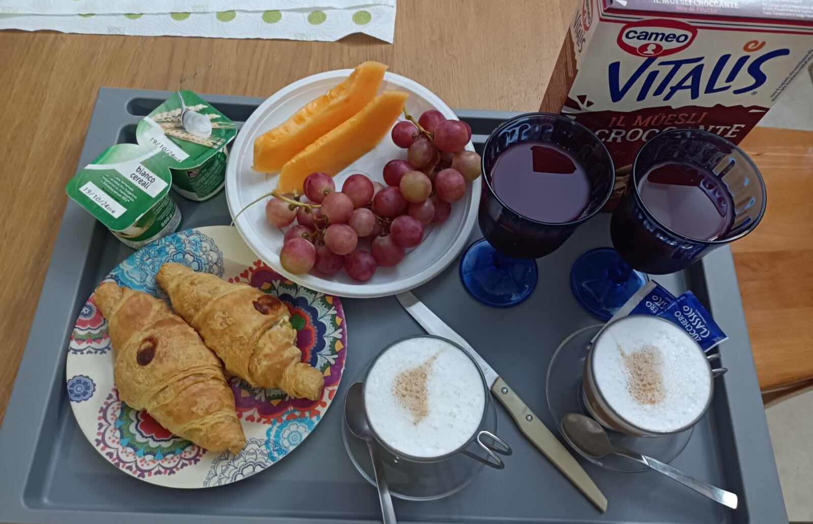 A breakfast tray with croissants, grapes, orange slices, cereal, two glasses of juice, and a box of Vitalis chocolate.