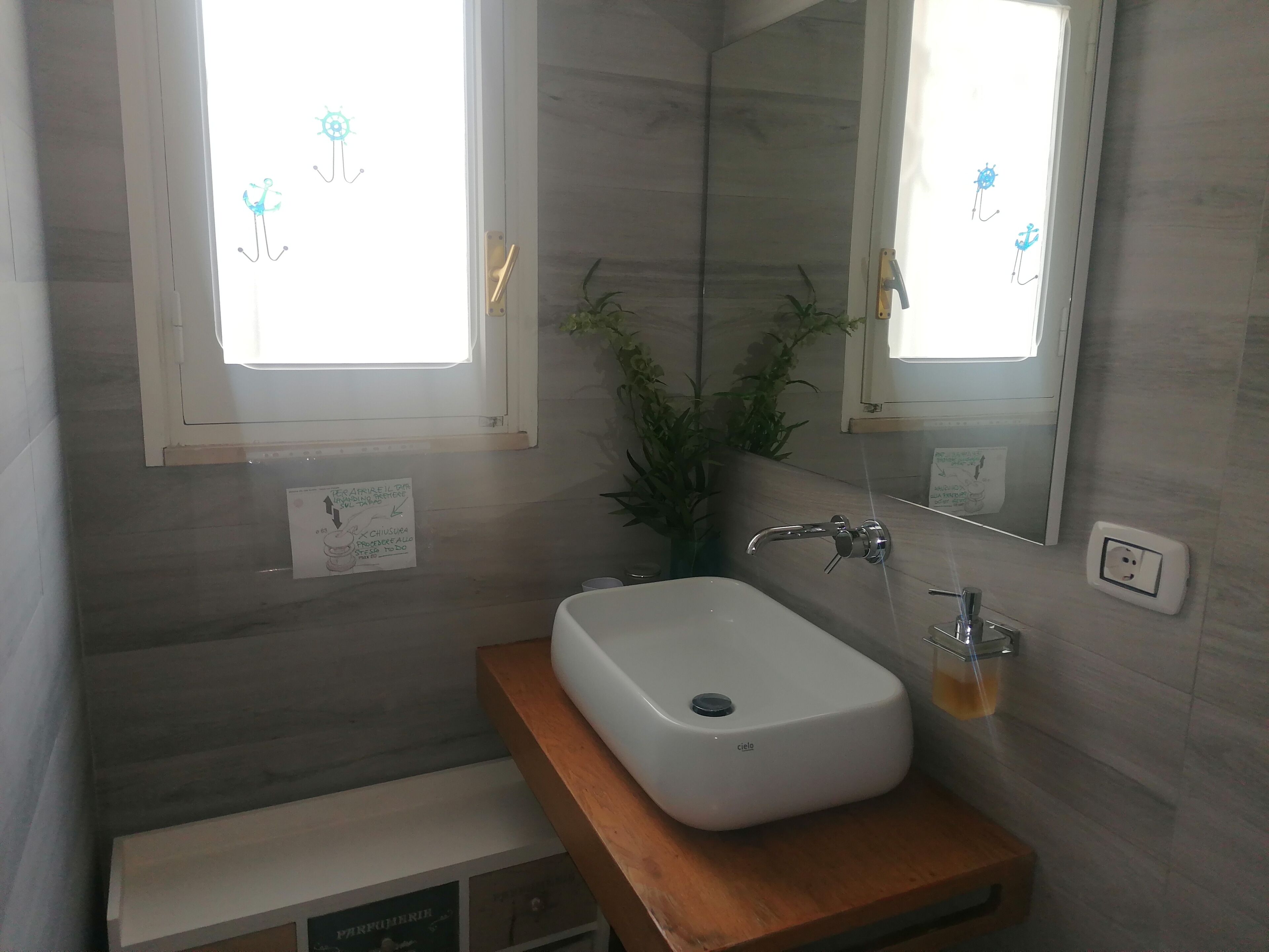 A bathroom with a white sink, wooden countertop, and a mirror above it. There is a window with a view of a ship and a sign on the wall.