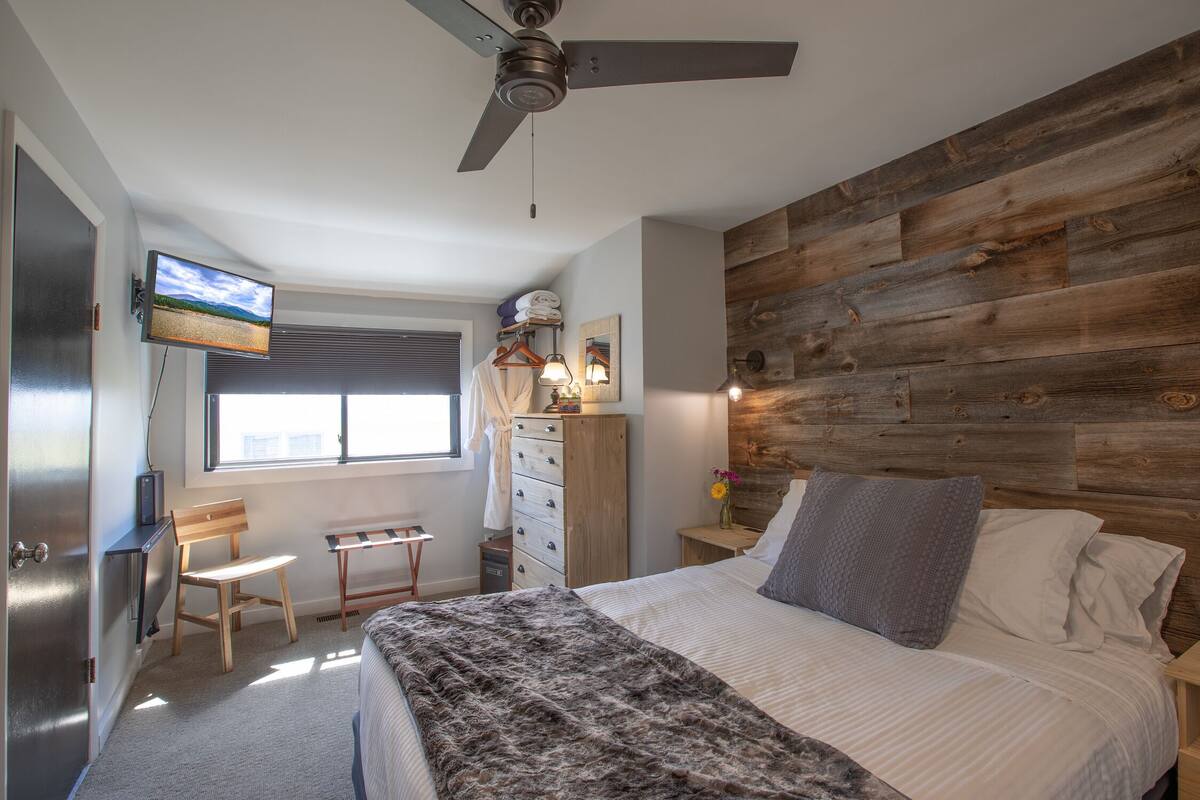 A bedroom with a bed, a desk, a chair, a TV mounted on the wall, and a wooden headboard.
