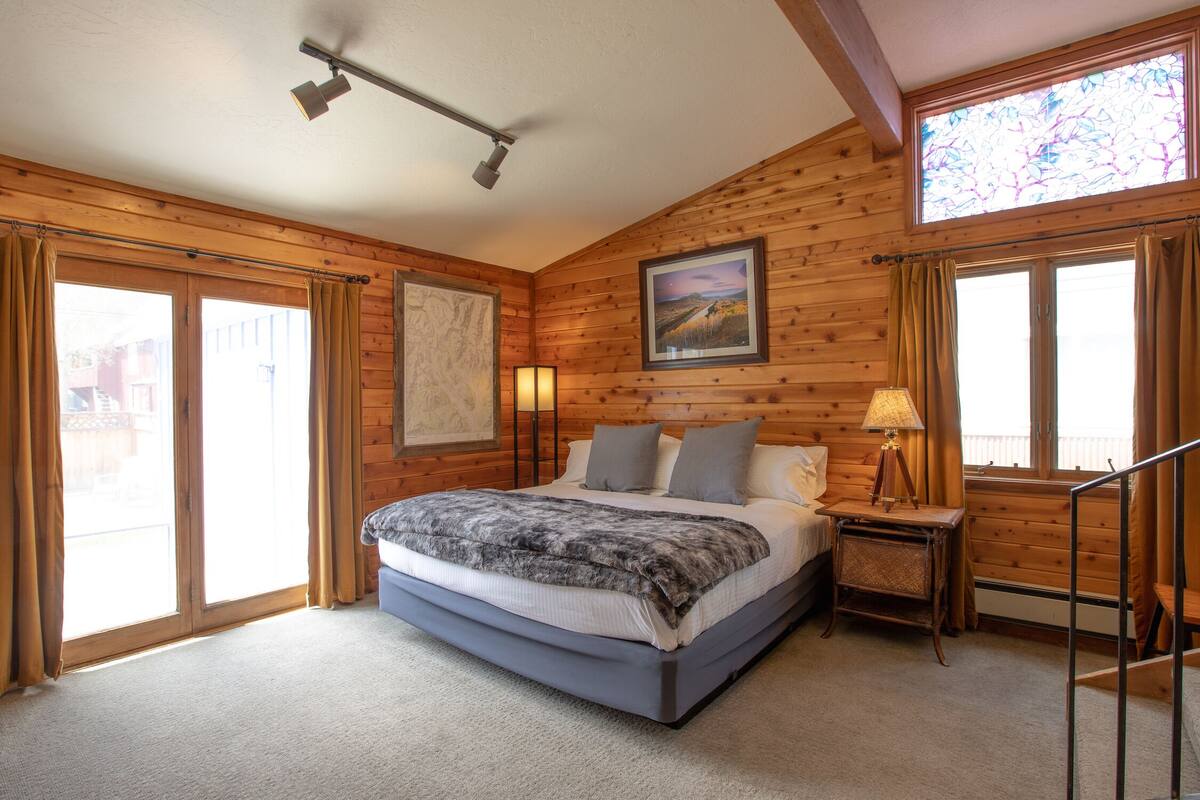 A bedroom with a large bed, wooden walls, a window with curtains, a nightstand with a lamp, and a painting on the wall.