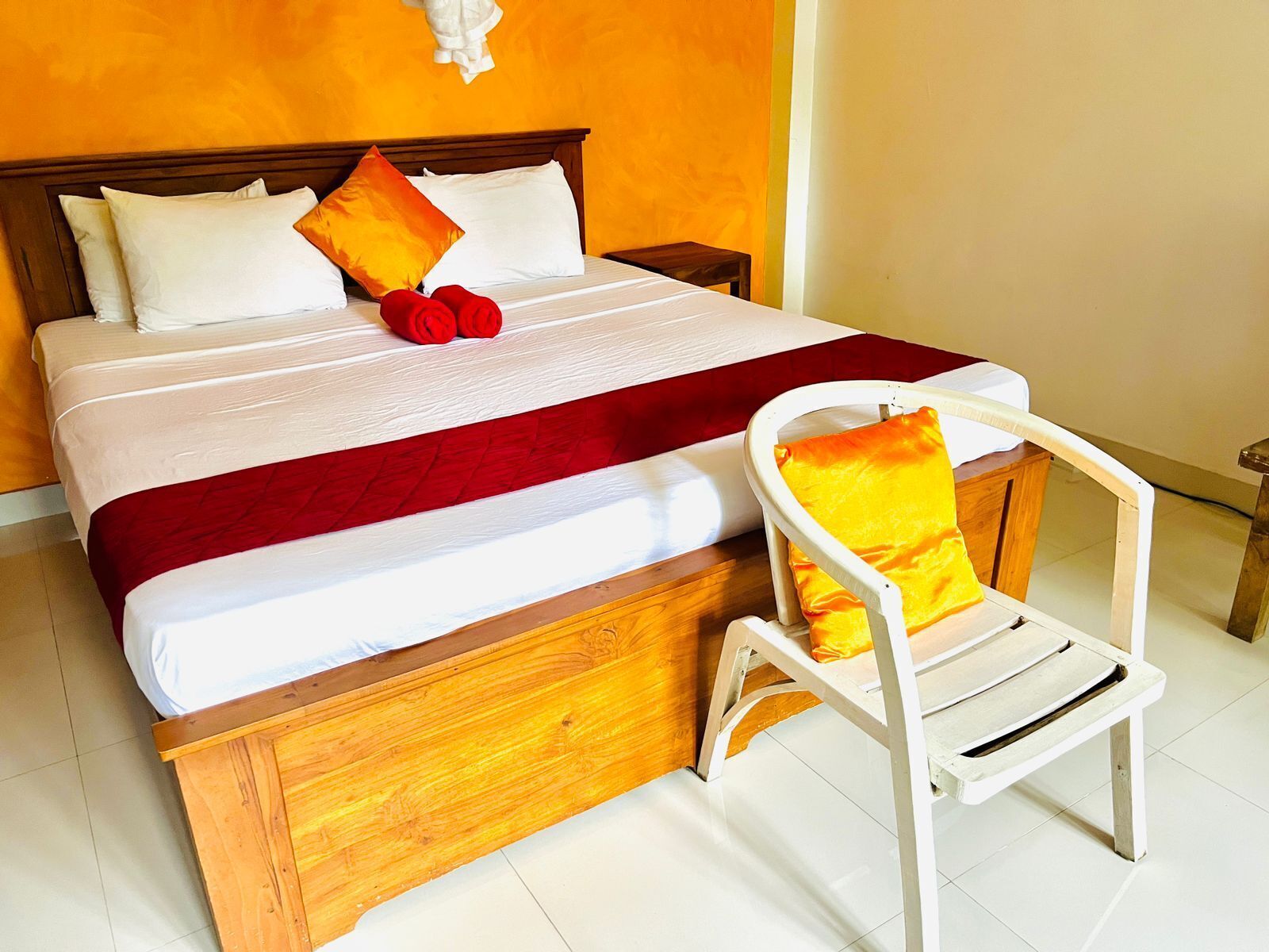 Una cama con cabecera de madera, ropa de cama blanca y almohadas rojas y amarillas. Una silla blanca con cojín amarillo está al lado de la cama.