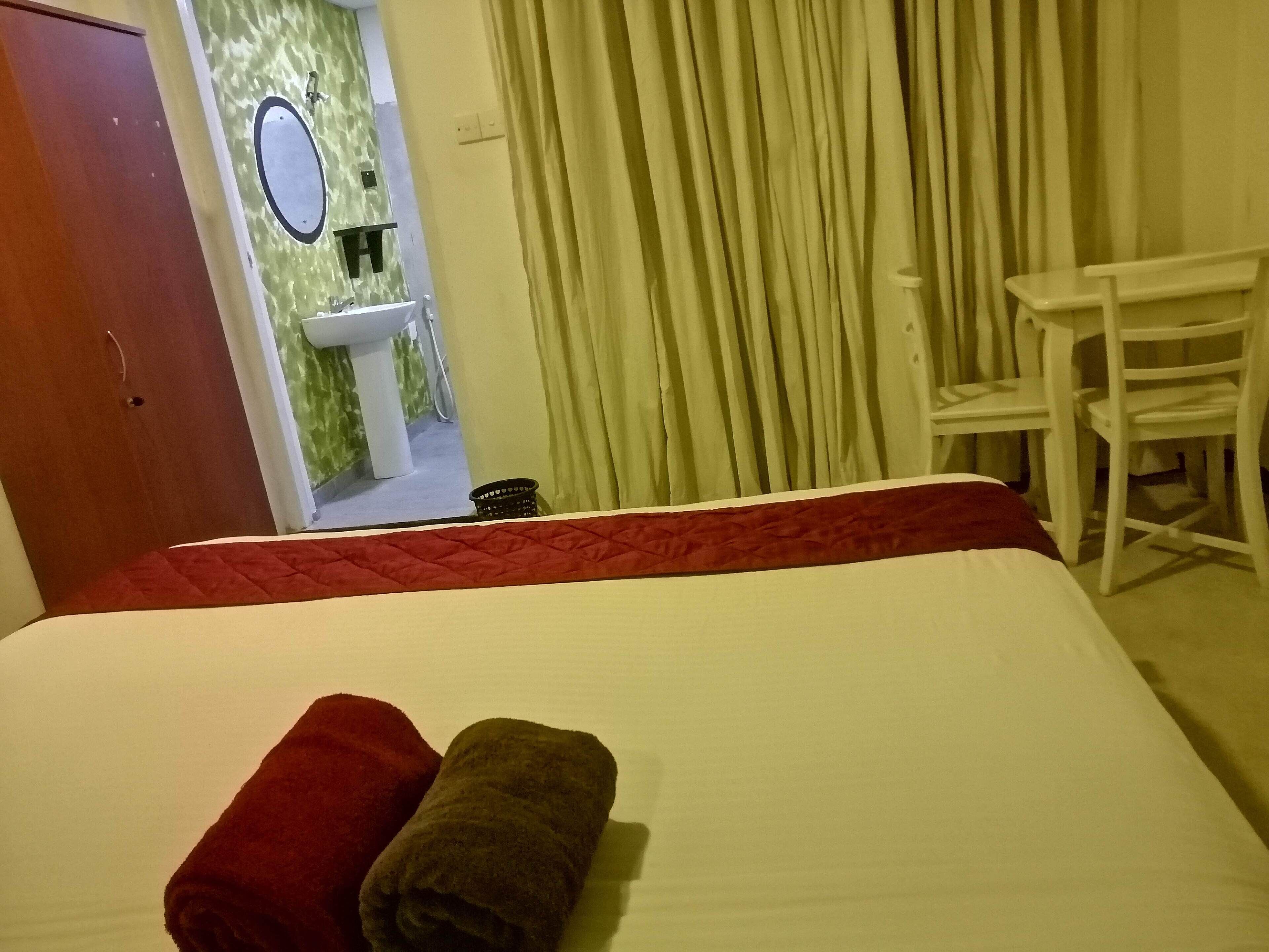 A hotel room with a bed, a red and brown towel, a white chair, a sink, and a mirror.