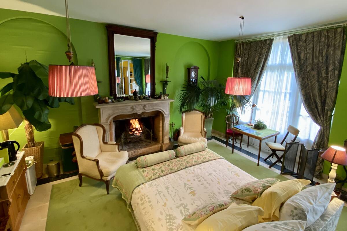 A room with a fireplace, green walls, a bed with floral bedding, a chair, a table, and a lamp.