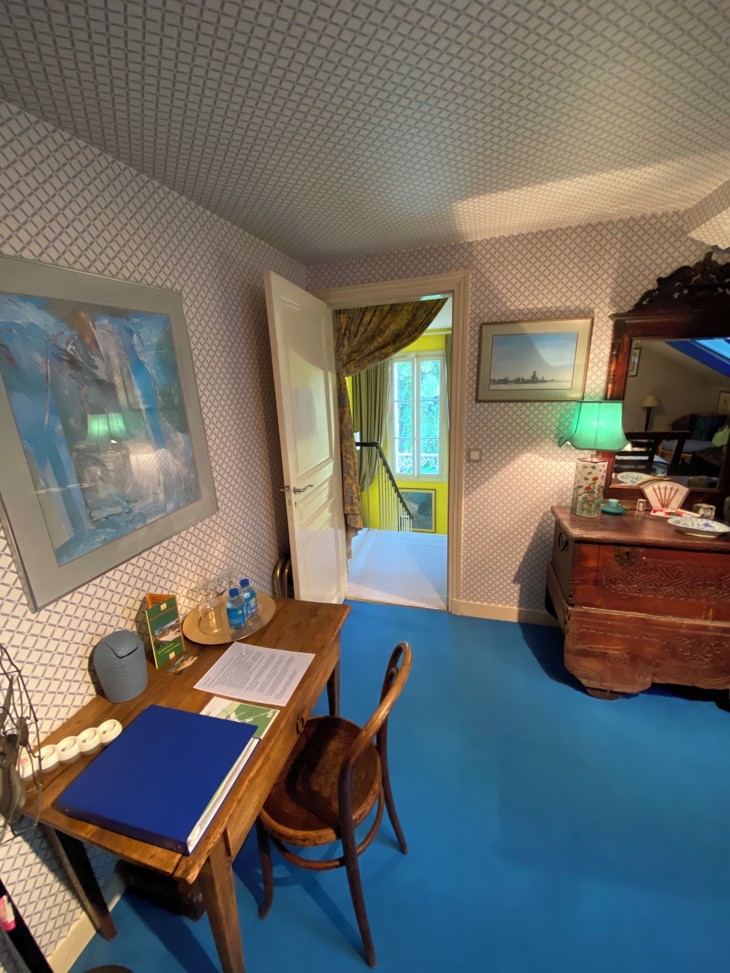 Una habitación con un escritorio de madera, una silla y una alfombra azul. Se ve una escalera que sube a un balcón con barandilla amarilla.