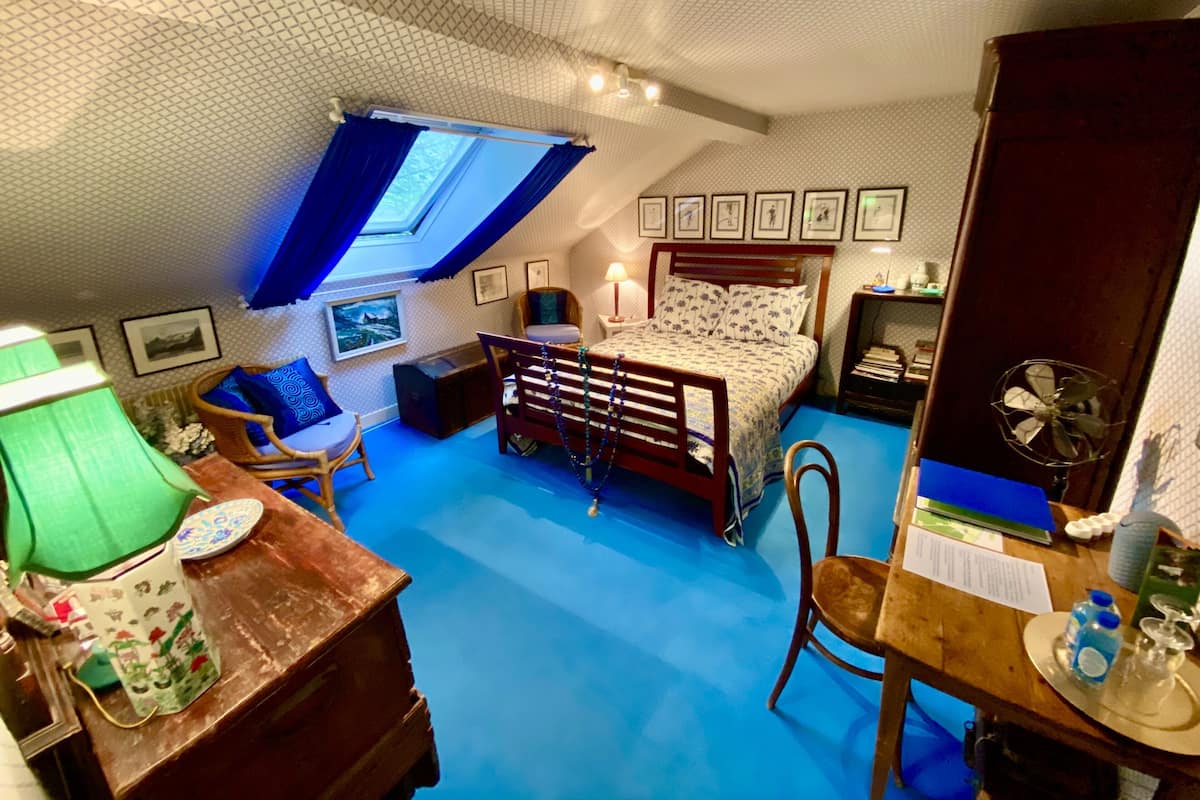 A bedroom with a sloped ceiling, a bed, a desk, a chair, and a window with blue curtains.