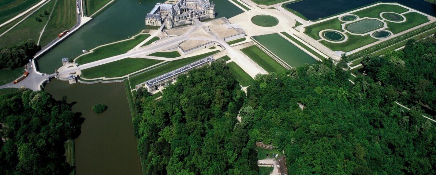 Vista aérea de una extensa propiedad histórica con jardines formales, un edificio central y un lago.