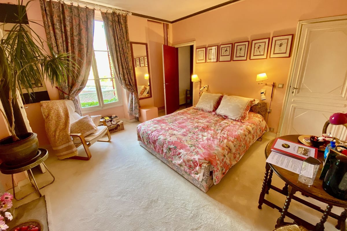 A bedroom with a floral bedspread, a potted plant, a chair, a bedside table with a lamp, and framed pictures on the wall.