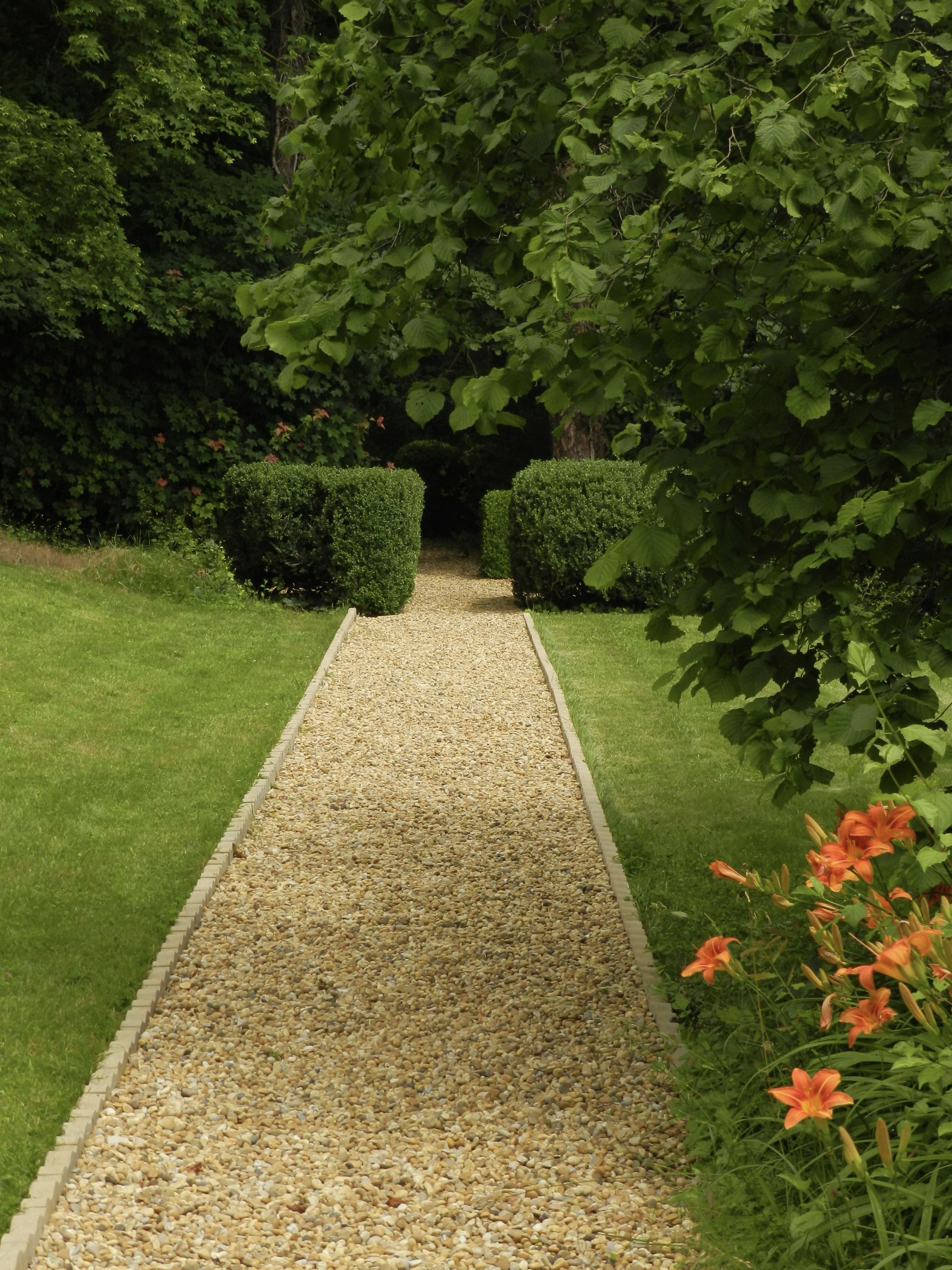 A garden pathway with trimmed hedges on either side, leading to a shaded area.