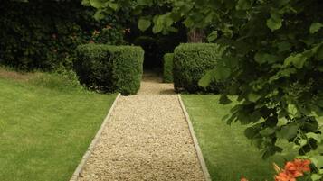A garden pathway with trimmed hedges on either side, leading to a shaded area.