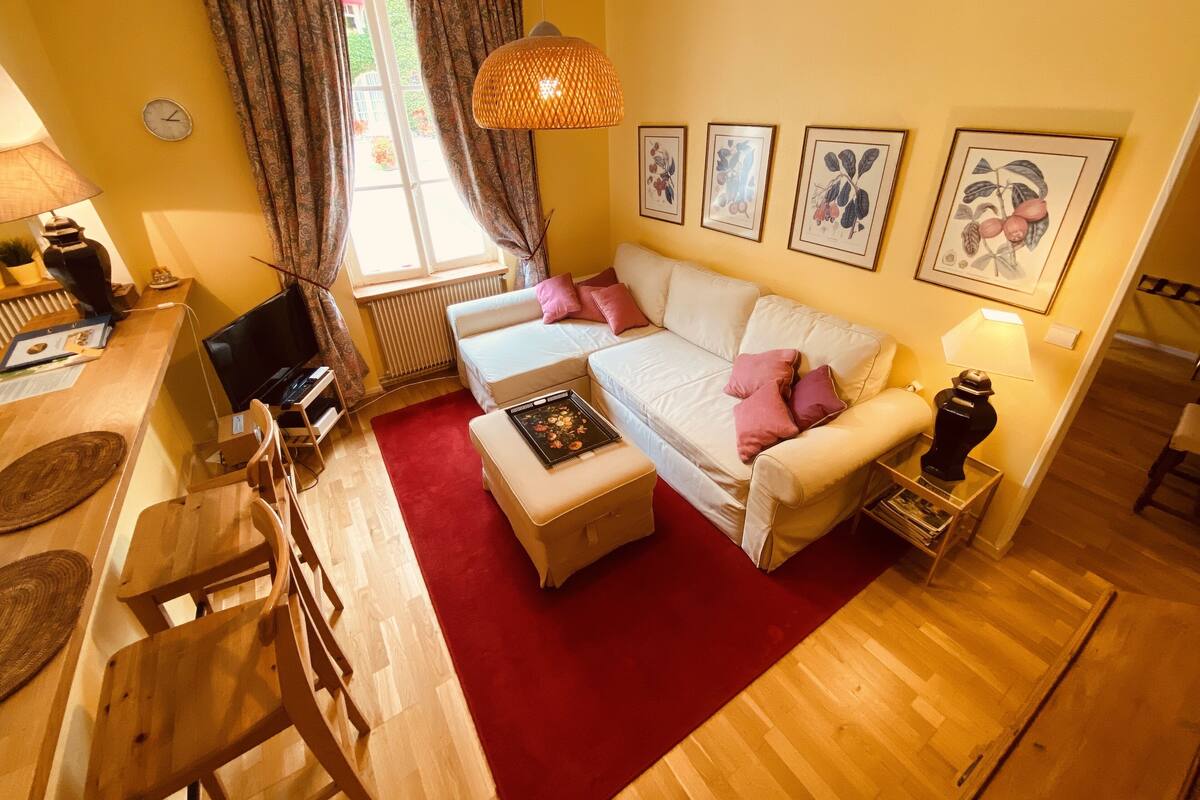 A cozy living room with a sofa, a coffee table, and a TV. There is a window with curtains, a lamp, and framed pictures on the wall.